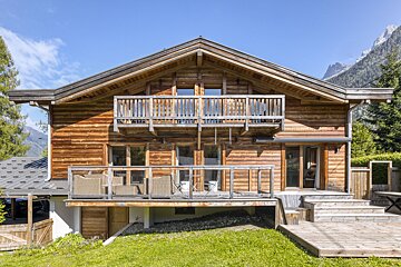 A multi-story wooden chalet with two balconies, a deck, and green lawn, nestled under a clear sky with mountains in the background.