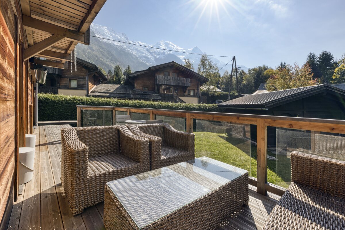 A sunlit wooden deck with wicker furniture overlooks a lush lawn, wooden chalets, and majestic snow-capped mountains under a bright sky.