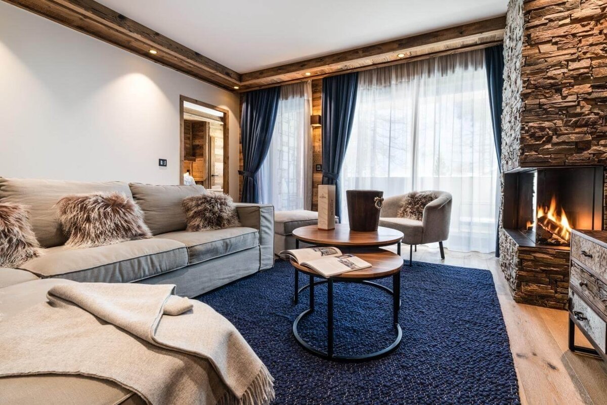 A cozy, rustic modern living room features a lit stone fireplace, plush sofa, dark blue rug, wooden accents, and large windows.