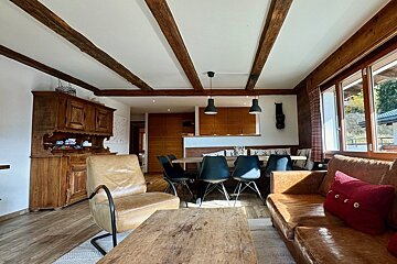 Rustic chalet interior with wooden beams, leather sofa & chair, dining area with dark chairs & table, antique hutch, and bright windows.