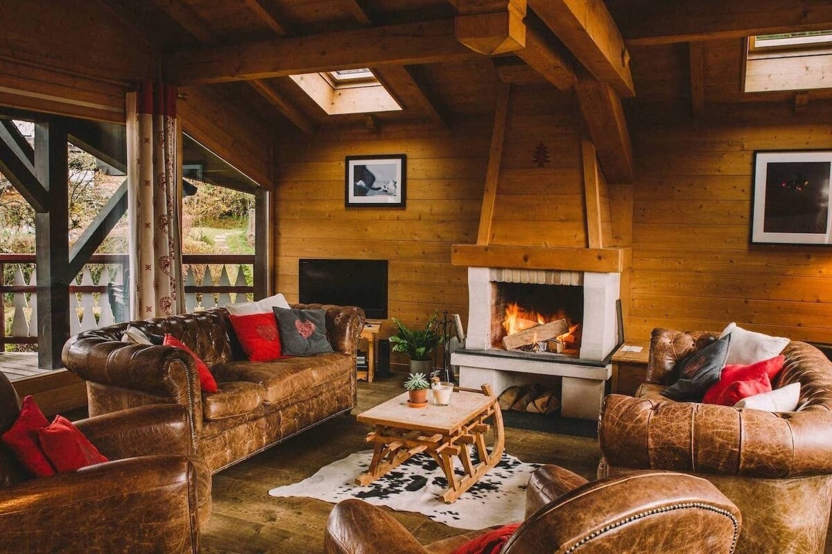 An inviting chalet living room with plush leather seating, a roaring fireplace, warm wood paneling, and a unique sled coffee table.
