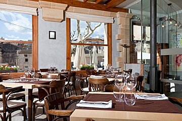 An inviting restaurant dining area with set tables, wine glasses, and expansive windows offering a scenic view of outdoor trees and buildings.