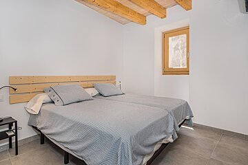 A cozy bedroom with two beds, striped bedding, and a wooden headboard. It features exposed wooden ceiling beams, white walls, a window, and tiled floor.
