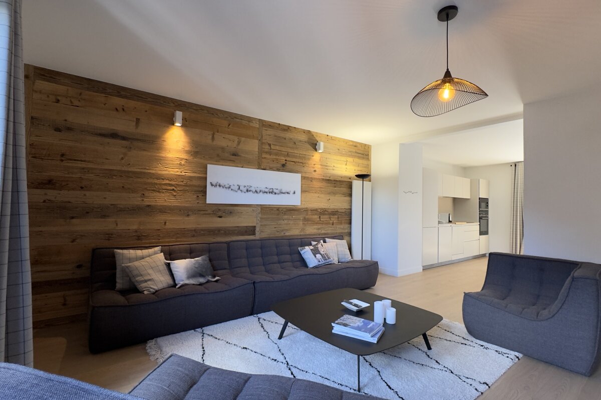 Stylish living room with a striking wood accent wall, modern gray sofa, black coffee table, and white rug. An open-plan kitchen is in the background.