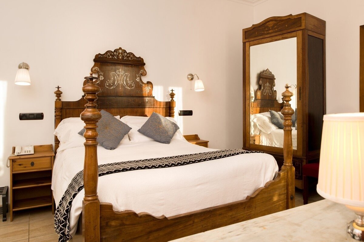 A cozy bedroom features an ornate wooden four-poster bed with white bedding, grey pillows, and a mirrored wardrobe.