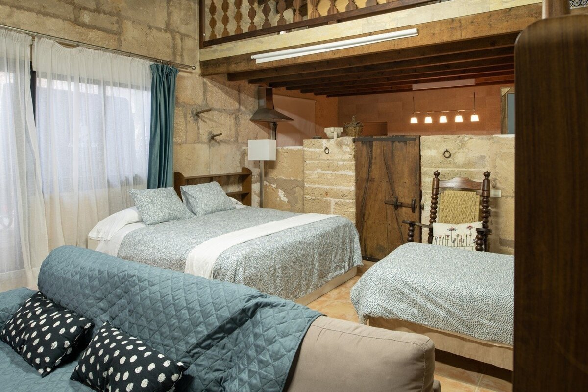 A cozy, rustic bedroom featuring stone walls, wooden beams, a large bed, a sofa, and a single bed. A wooden mezzanine overlooks the space.