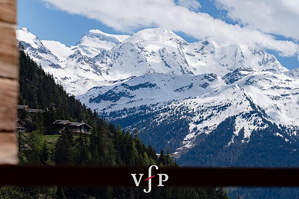 Majestic snow-capped mountains rise above a forested slope with chalets, under a blue sky with clouds. A wooden frame is on the left.