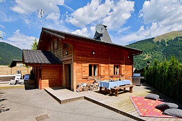 Cozy wooden chalet nestled in the mountains on a sunny day. Features an outdoor dining area, hot tub, and lush green surroundings.