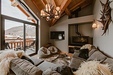 A cozy chalet living room features a fireplace, antler chandelier, and fur accents, with a large balcony offering snowy mountain and village views.