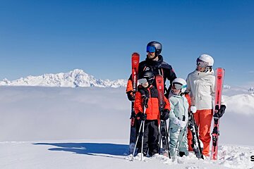 A family of four in ski gear stands on a snowy mountain summit above a sea of clouds under a clear blue sky.