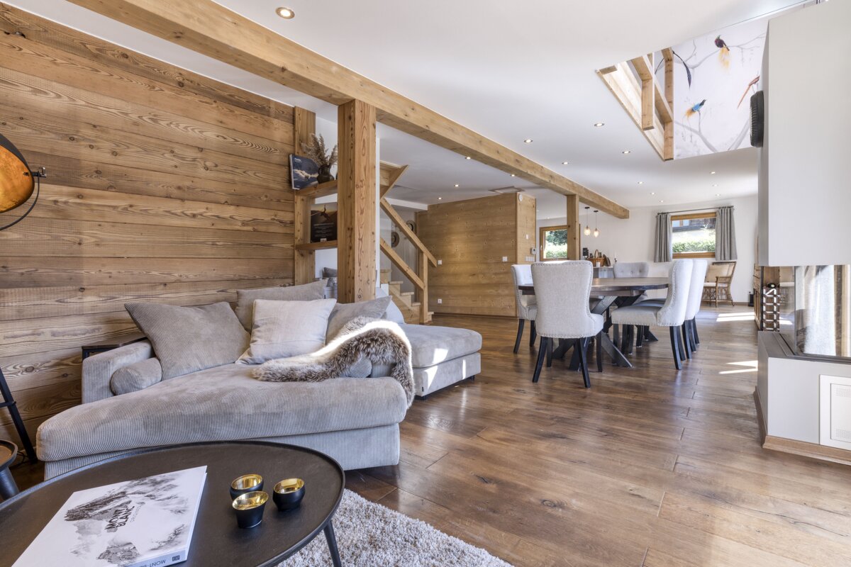 A cozy, modern chalet-style living and dining room with wood-paneled walls, exposed beams, an L-shaped sofa, dining table, and fireplace.