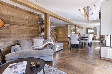 A cozy, modern chalet-style living and dining room with wood-paneled walls, exposed beams, an L-shaped sofa, dining table, and fireplace.