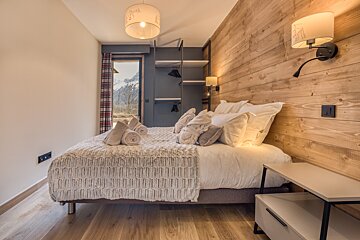 A cozy chalet-style bedroom with a bed, wood paneling, open wardrobe, and a window framing a snowy mountain view. Warm lighting enhances the inviting atmosphere.