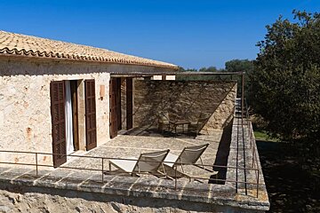 Sunny stone house with a tiled roof & balcony featuring lounge chairs and shaded seating, under a bright blue sky.