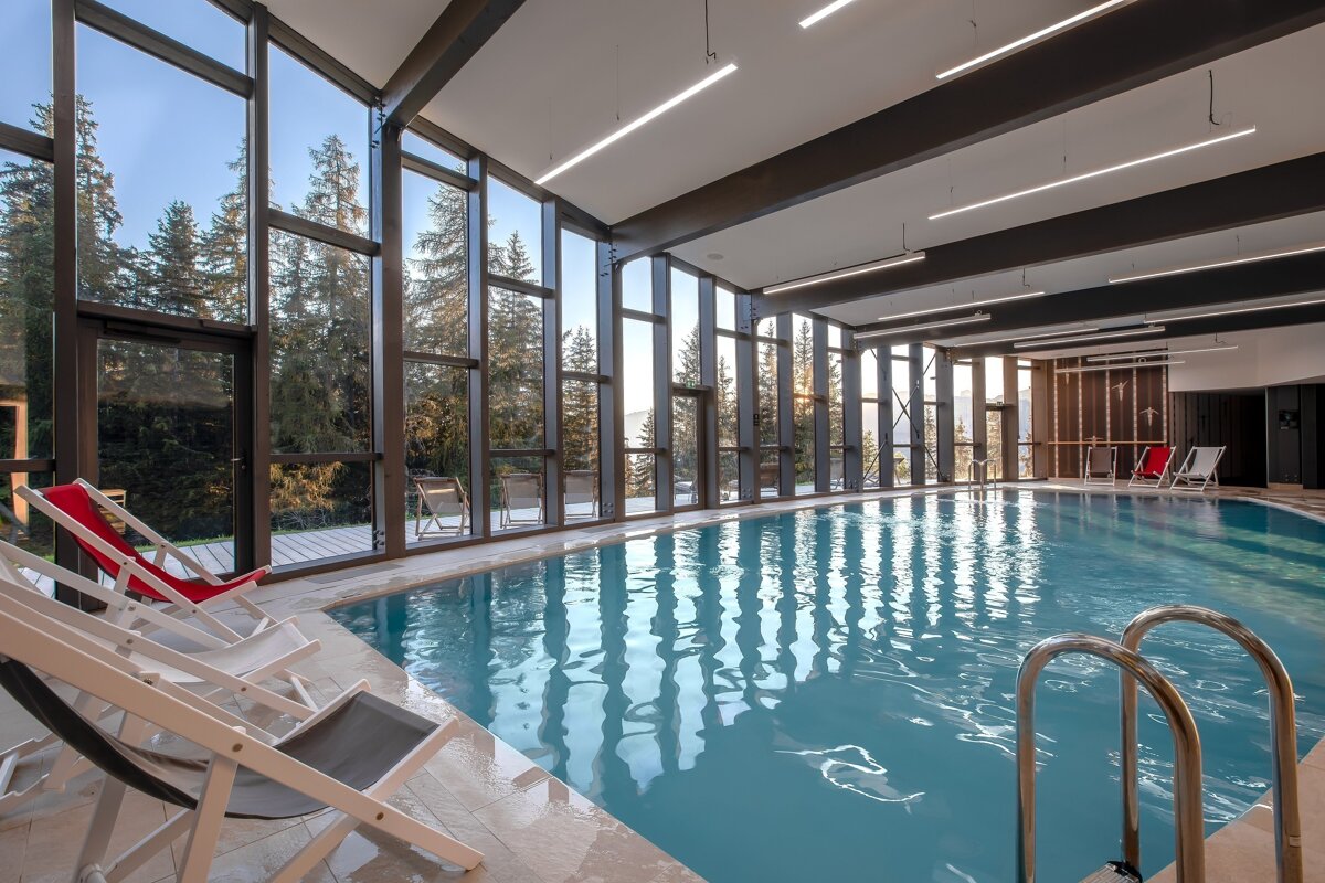 A modern indoor pool with expansive windows showcases a lush pine forest and mountain landscape. Loungers are arranged poolside for relaxation.