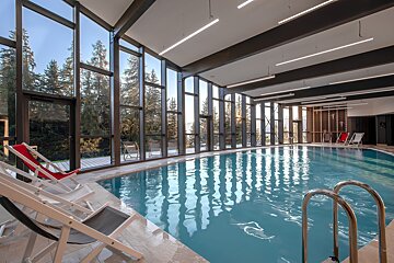 A modern indoor pool with expansive windows showcases a lush pine forest and mountain landscape. Loungers are arranged poolside for relaxation.