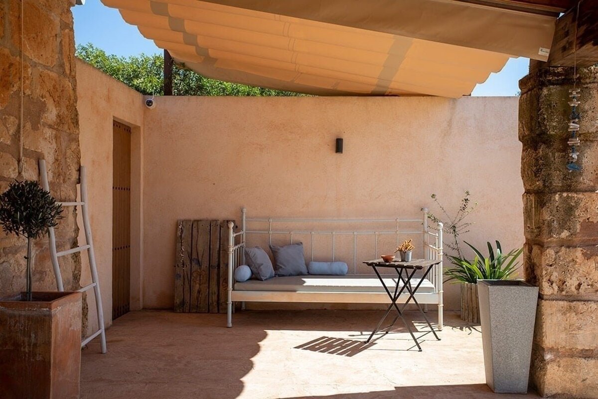 A sun-drenched, rustic outdoor patio with a comfy white daybed, small table, and a retractable awning. Stone and peach-toned walls create a peaceful retreat.