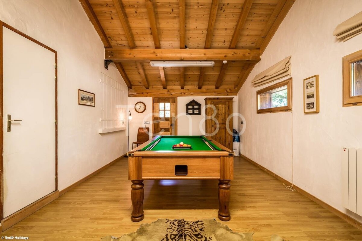 A rustic game room features a central pool table, exposed wooden ceiling beams, light wood floors, and white walls with windows.