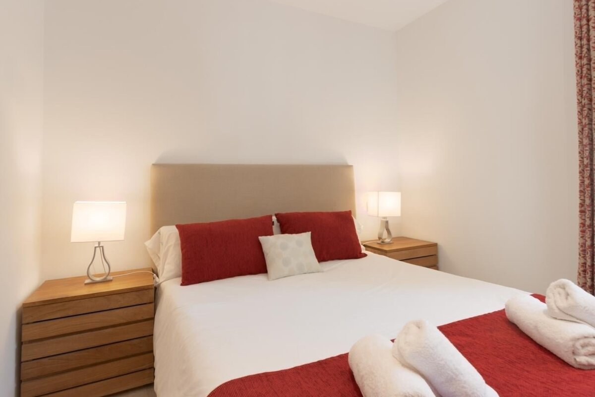 A simple bedroom with a neatly made bed, white sheets, red pillows, and a red runner. Two nightstands with lamps flank the bed.