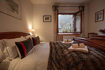 A cozy bedroom featuring a large bed with a faux fur throw, an open window looking out to an autumn mountain village, and warm, inviting lighting.