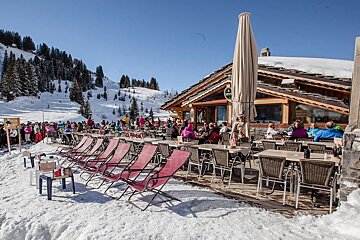 A bustling ski lodge patio on a sunny, snowy mountain with people dining, red lounge chairs, and a rustic wooden building under a clear blue sky.