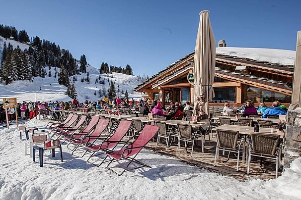 A bustling ski lodge patio on a sunny, snowy mountain with people dining, red lounge chairs, and a rustic wooden building under a clear blue sky.