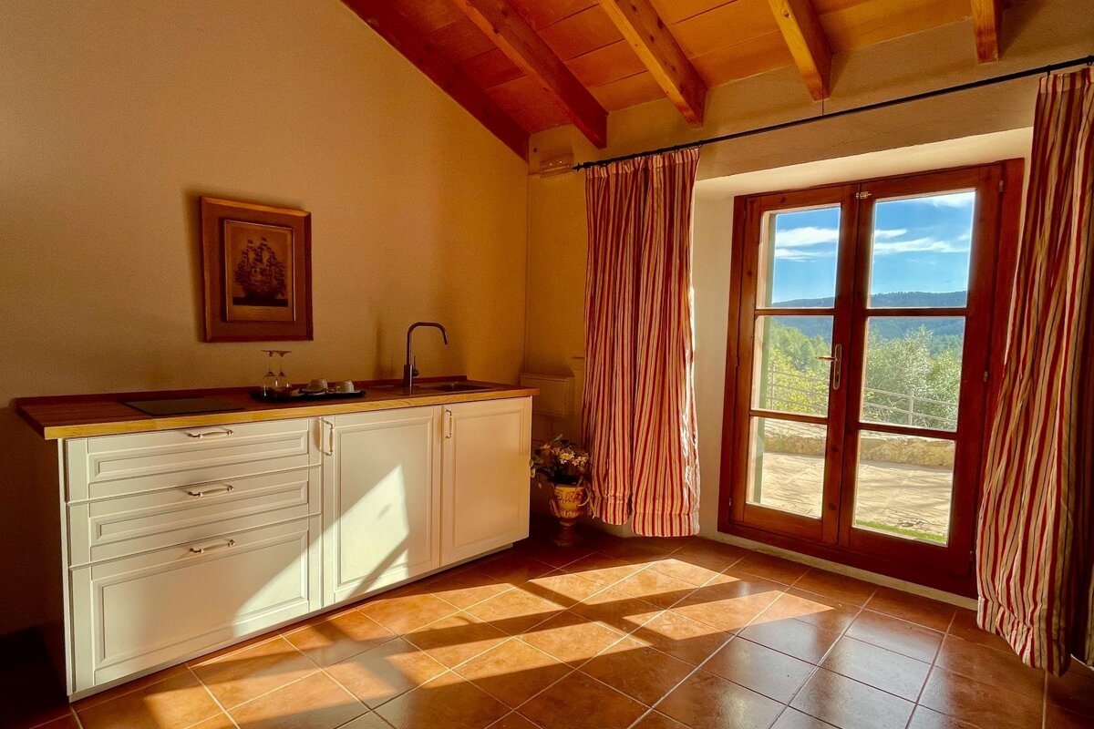 A sun-drenched room with a kitchenette, exposed wood ceiling, terracotta tiles, and a window framing a lush, green view. Red curtains.