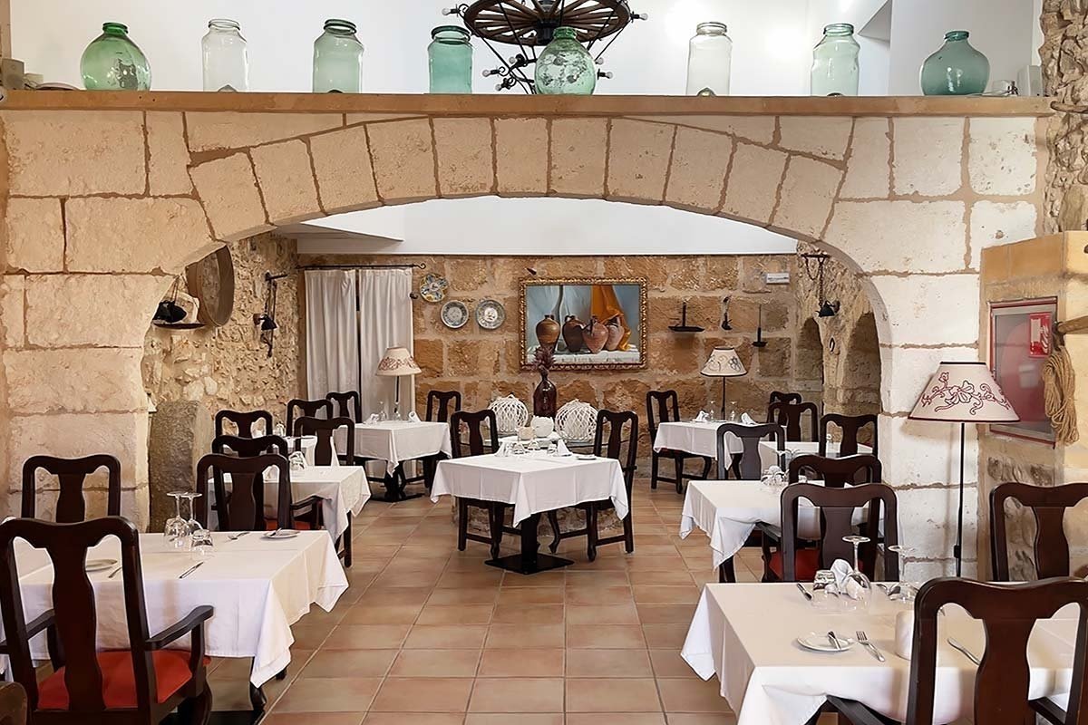 Rustic restaurant interior with stone walls, archways, and neatly set tables with white tablecloths. Decorative glass jars adorn an upper shelf.