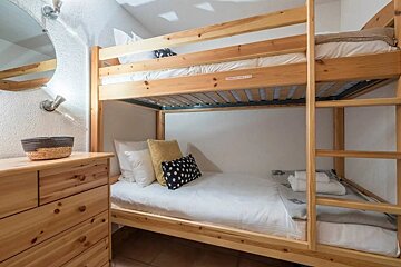 A cozy bedroom features light wooden bunk beds with white bedding, accent pillows, a matching dresser, and a round mirror with wall lights.
