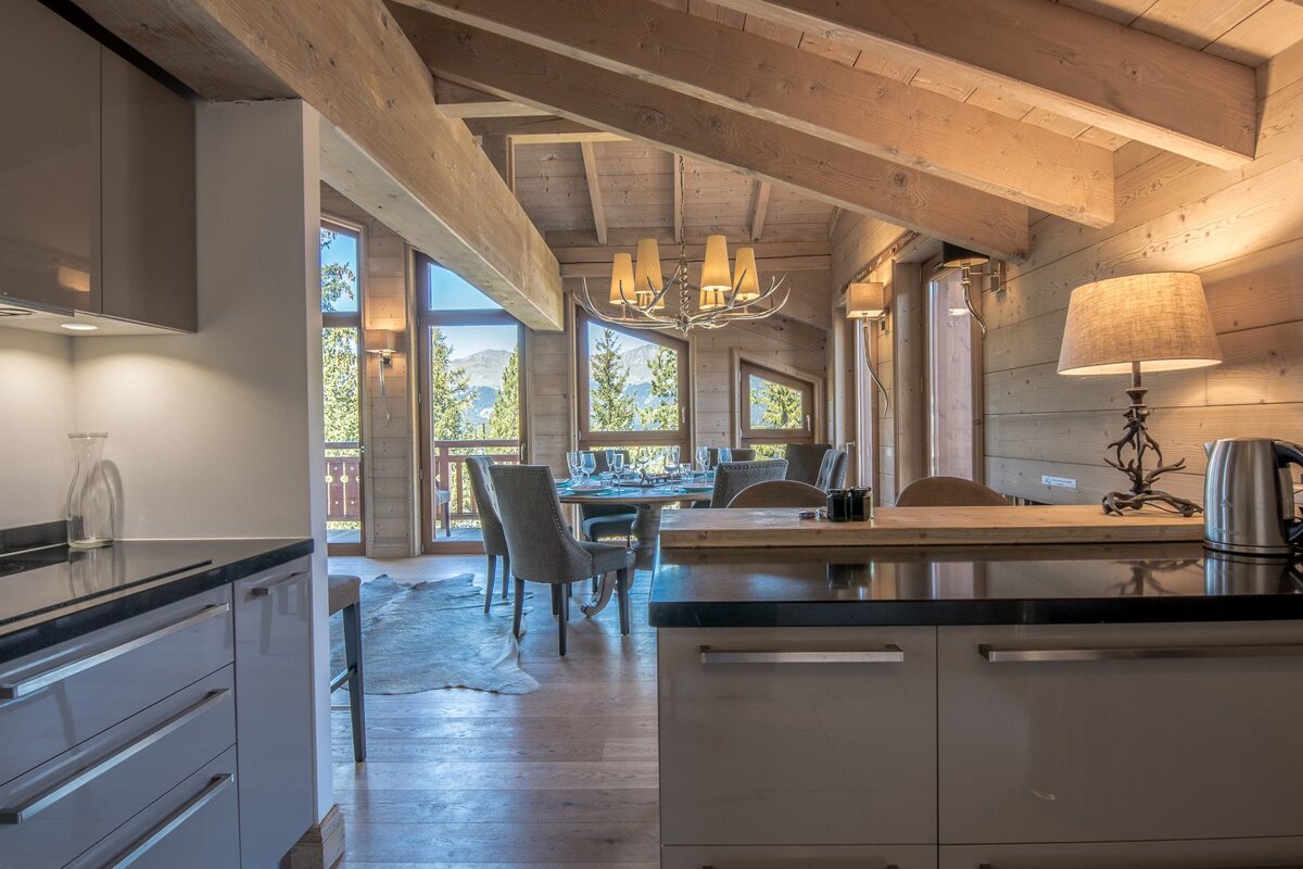 A modern kitchen overlooks a rustic, wood-paneled dining room with exposed beams, large windows offering picturesque mountain views, and cozy lighting.