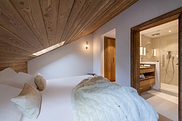 A modern attic bedroom features a sloped wooden ceiling, white bed with plush blanket, and an open ensuite bathroom with a shower and vanity.