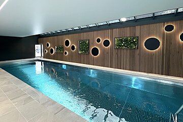 A modern indoor swimming pool with blue water, a wooden slatted wall featuring backlit circular lights and framed plant art, under skylights.