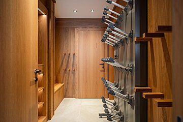 A modern, wood-paneled ski room featuring a multi-tiered boot drying rack, a long bench, and subtle built-in lighting.