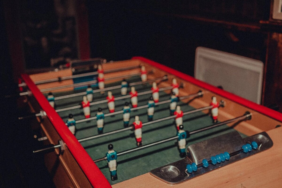 A dimly lit foosball table with red and blue players on a green field. A score counter with blue beads is visible.