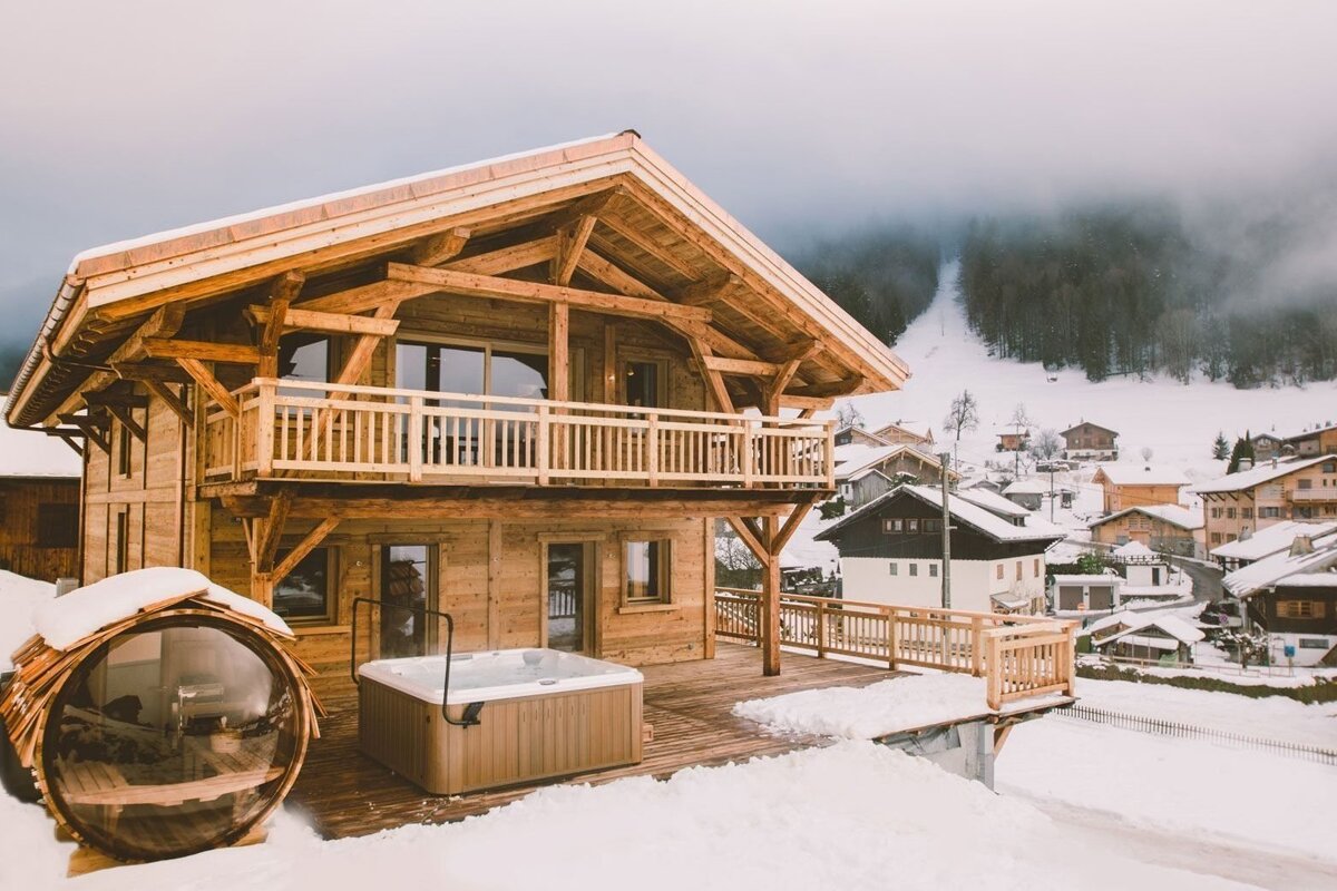A cozy wooden chalet with a jacuzzi and barrel sauna on a snowy deck, overlooking a charming mountain village and a forest ski slope under a grey sky.