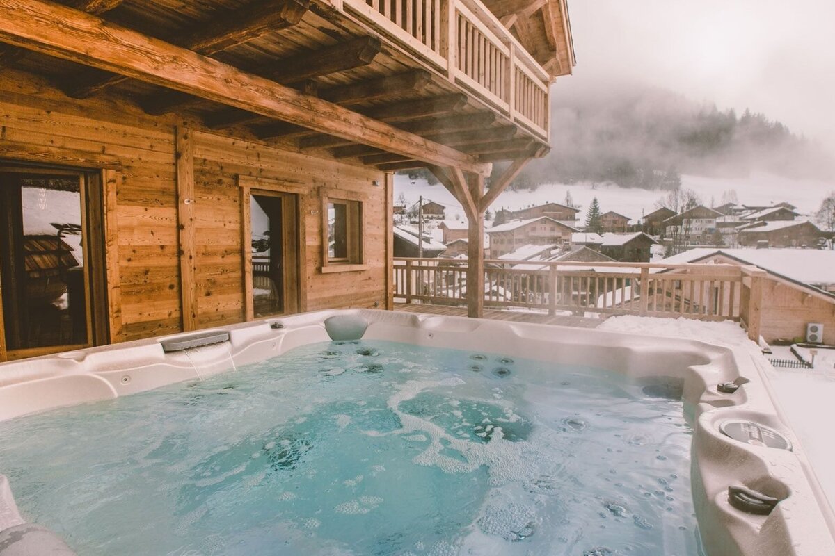 An outdoor hot tub on a wooden chalet's deck, overlooking a snowy mountain village. Steam rises from the hot, bubbling water in the winter landscape.