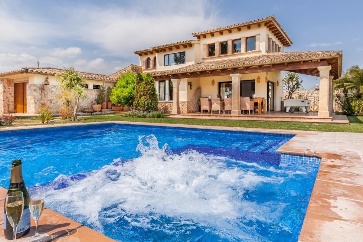 A sunny villa with a large swimming pool, including a bubbling jacuzzi. A bottle of champagne and two glasses sit poolside, with an outdoor patio in the background.