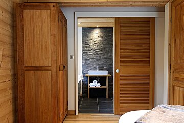 A rustic wooden room with a large wardrobe, looking into a modern bathroom with a stacked stone wall, white sink on a wooden vanity, and dark floor tiles.