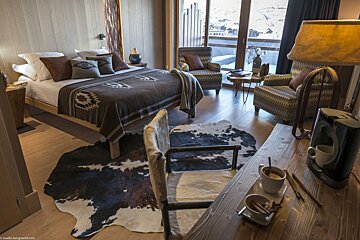 A cozy, rustic bedroom features a bed with a patterned blanket, a cowhide rug, armchairs by a snowy window, and a desk with coffee.
