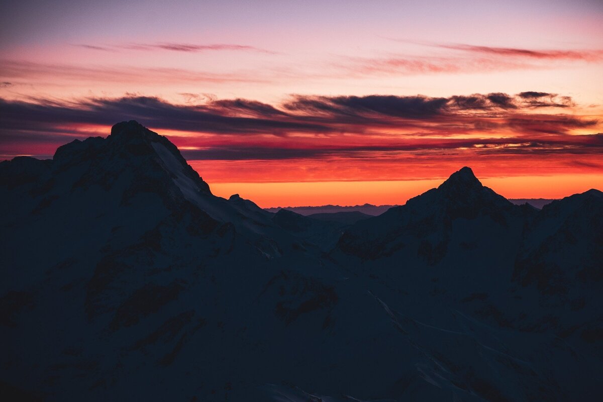Silhouetted mountain peaks frame a vibrant sunset sky. Fiery orange and red colors blend into soft purple, with dramatic clouds overhead.