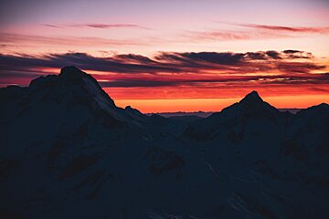 Silhouetted mountain peaks frame a vibrant sunset sky. Fiery orange and red colors blend into soft purple, with dramatic clouds overhead.