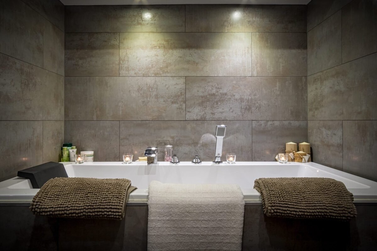 A cozy, modern corner bathtub with textured grey walls, soft uplighting, and glowing candles, featuring plush towels for a relaxing, spa-like atmosphere.