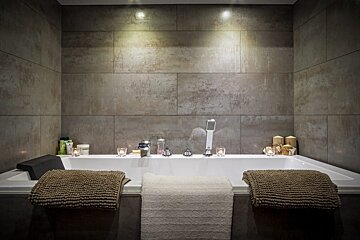 A cozy, modern corner bathtub with textured grey walls, soft uplighting, and glowing candles, featuring plush towels for a relaxing, spa-like atmosphere.