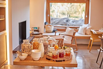 A bright, modern room features a breakfast buffet with cereals, milk, jams, and juices on a wooden table, overlooking a large window.