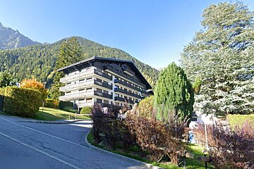 A large chalet-style apartment building nestled in wooded mountains, with lush trees and a paved road in the foreground under a clear blue sky.