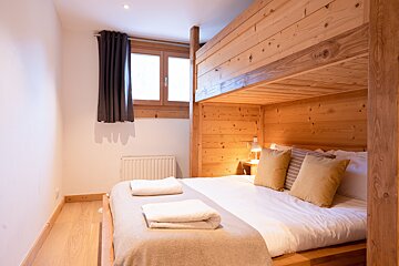 A cozy bedroom featuring a wooden bunk bed over a double bed with neutral bedding, a bright window with dark curtains, and light wooden flooring.