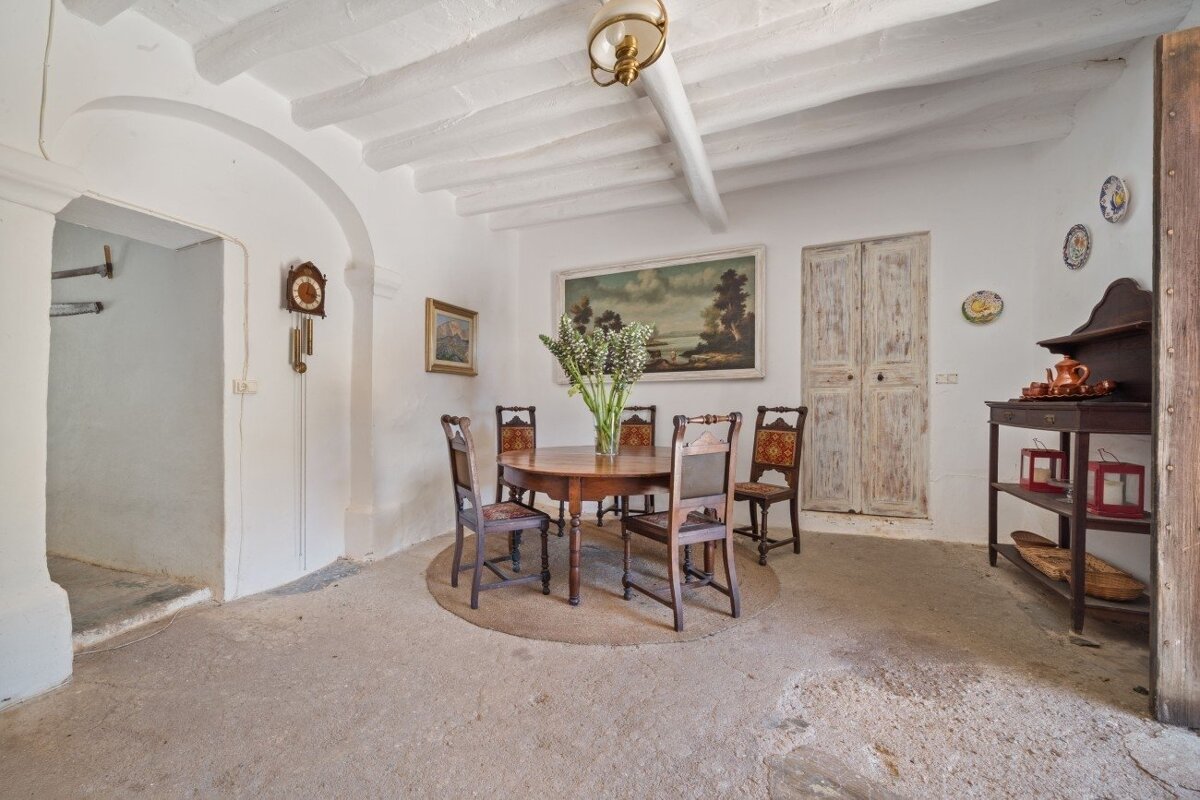 A rustic dining room with white walls, beamed ceiling, wooden table and chairs, antique clock, and traditional decor like paintings and old doors.