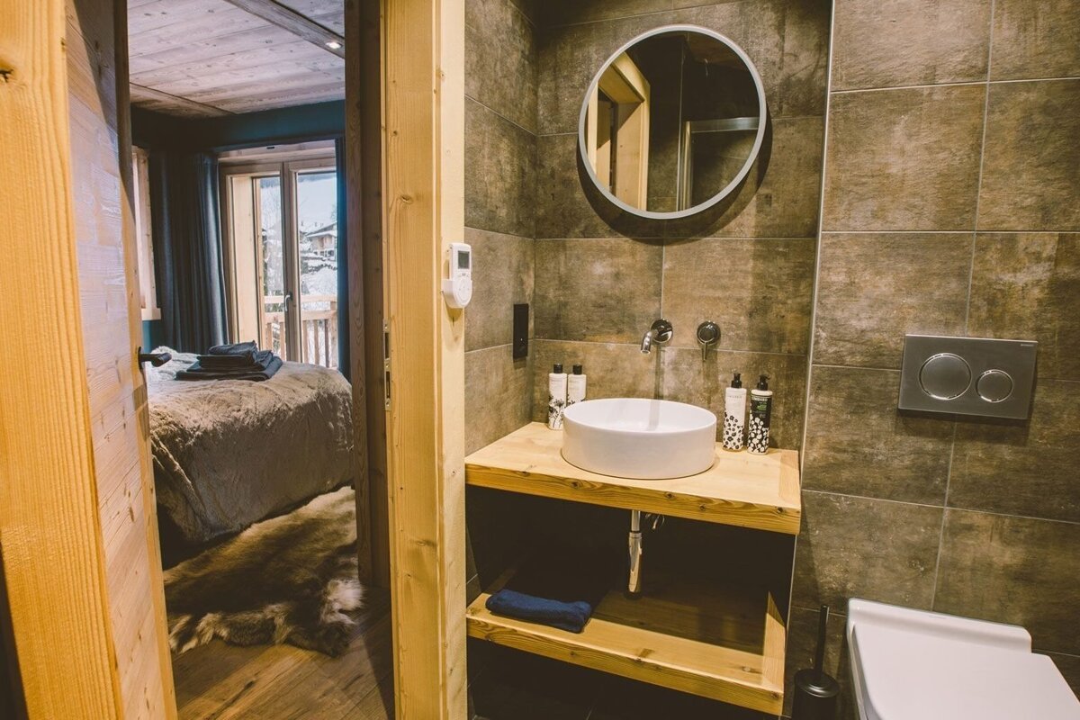 A modern bathroom with a wooden vanity, tiled walls, and a round mirror, adjacent to a cozy bedroom with a bed, fur rug, and balcony view.