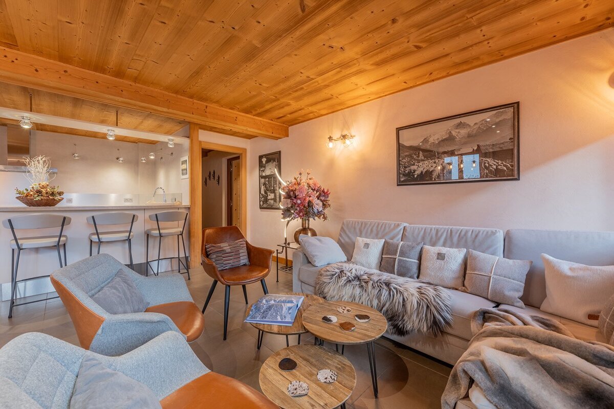 A cozy living area and adjacent kitchen bar featuring a wooden ceiling, comfy sofa with fur throws, stylish chairs, and mountain-themed art.