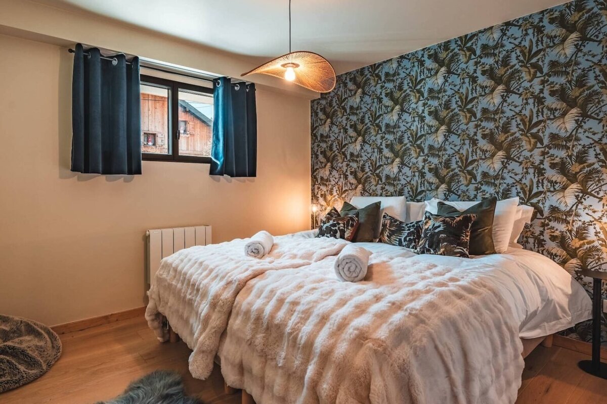 A cozy bedroom with a fluffy bed, tropical leaf accent wall, unique woven pendant light, and a window with blue curtains overlooking wooden buildings.